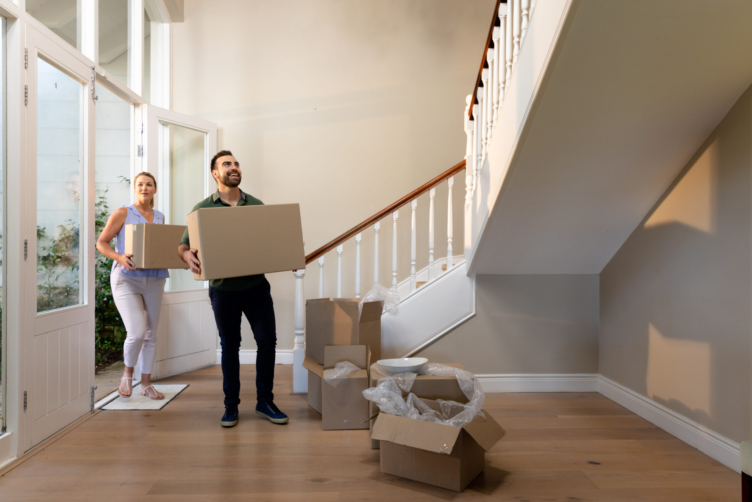 Homeowners arriving at their new home they purchased walking through the front door and standing in the hallway carrying cardboard packing boxes and looking around smiling, open packing boxes on the floor - Cal-West Property Inspections Homeowners arriving at their new home they purchased walking through the front door and standing in the hallway carrying cardboard packing boxes and looking around smiling, open packing boxes on the floor - Cal-West Property Inspections