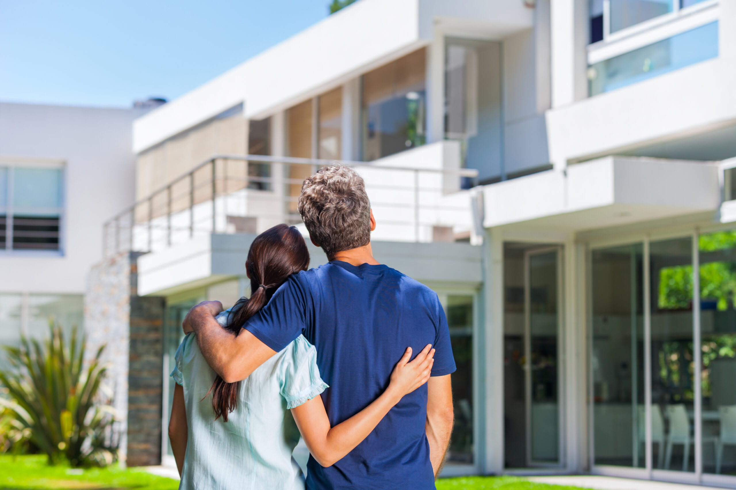 New homeowners embracing in front of new big modern house, looking at their new home - Cal-West Property Inspections New homeowners embracing in front of new big modern house, looking at their new home - Cal-West Property Inspections